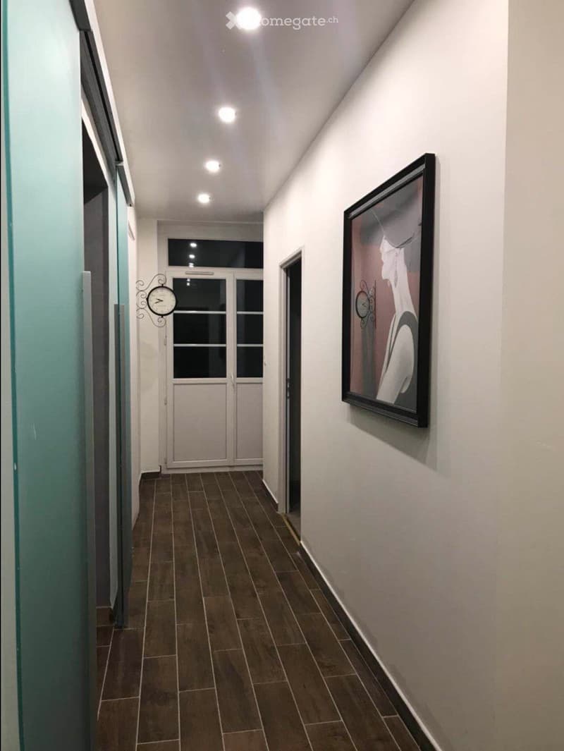 A narrow hallway with tiled flooring, white walls, and a framed artwork on the right side. A decorative wall clock is mounted on the left wall, and a double door with glass panels is visible at the end of the hallway.