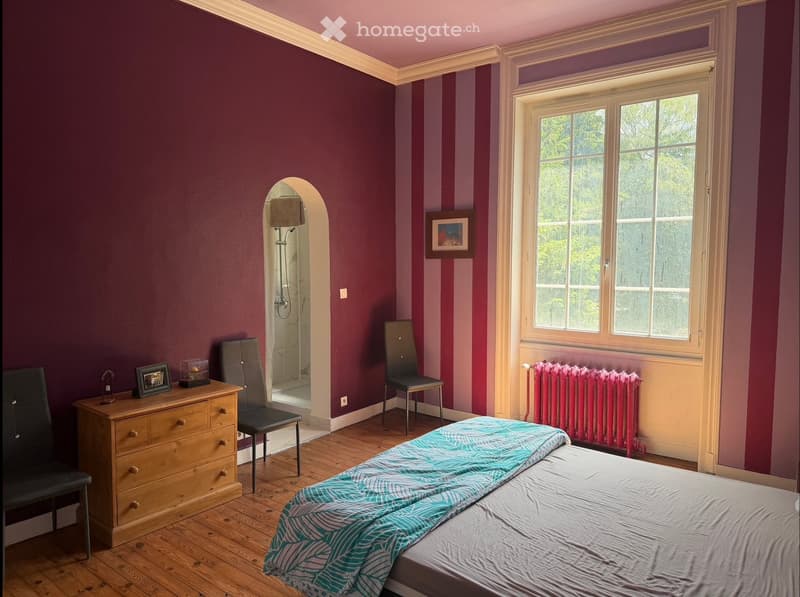 Bedroom with wooden floor, bed with teal and white patterned bedspread, two chairs, wooden dresser, radiator, picture frames, and a lamp.