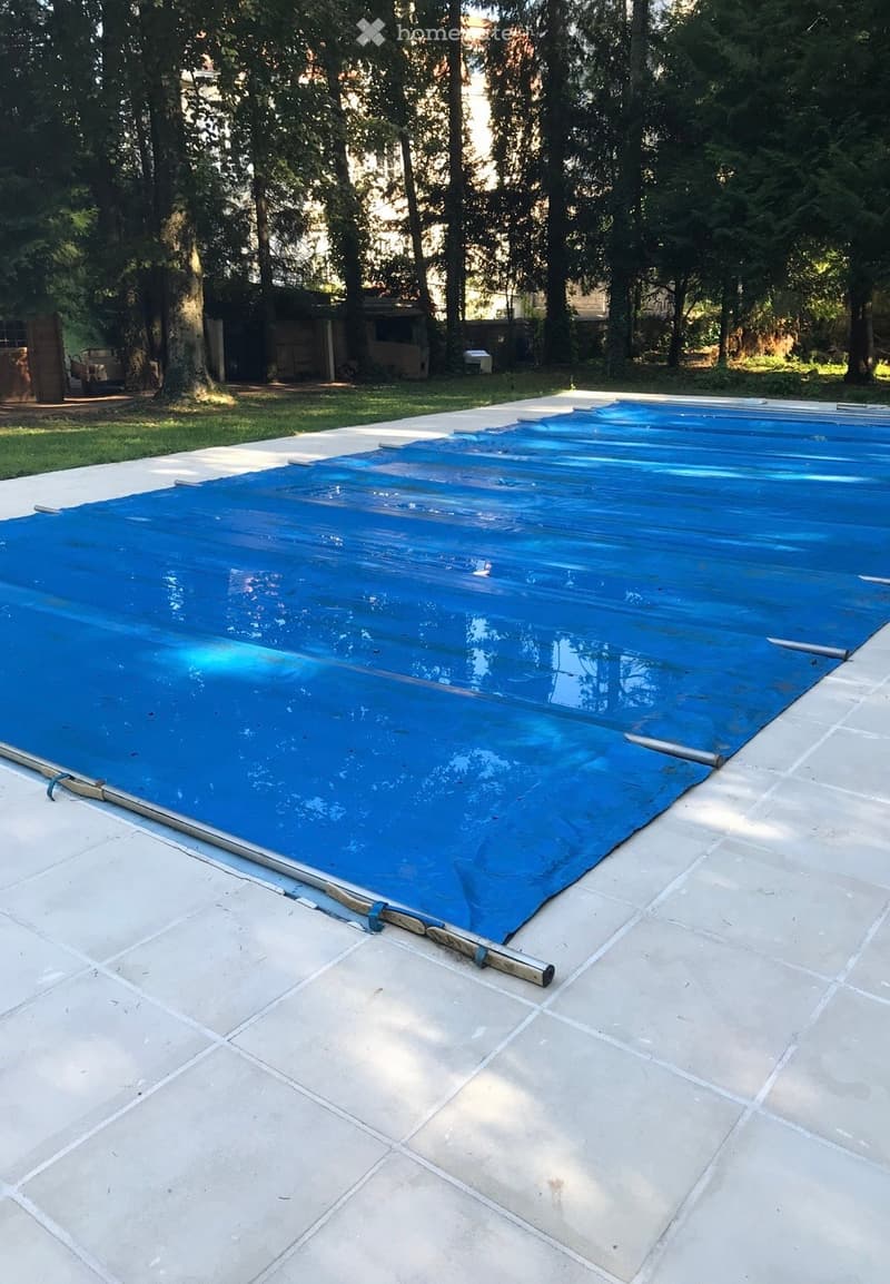 A swimming pool covered with a blue tarp, surrounded by a tiled patio and trees.