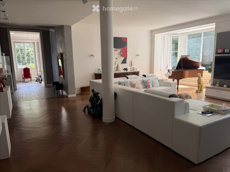 Spacious living room with wooden flooring, large white sectional sofa, colorful throw pillows, a grand piano, a television, and decorative statues.