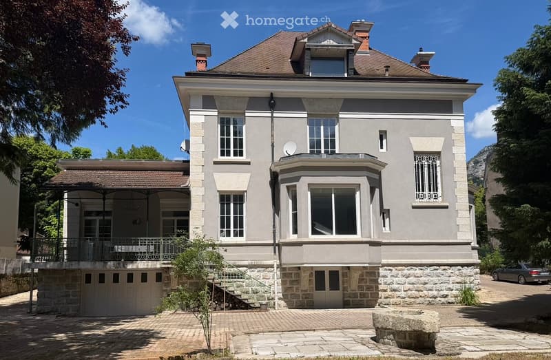 A two-story house with a gray facade, multiple windows, a balcony with metal railings, and a stone base. The house features a garage and is surrounded by greenery and a paved driveway.
