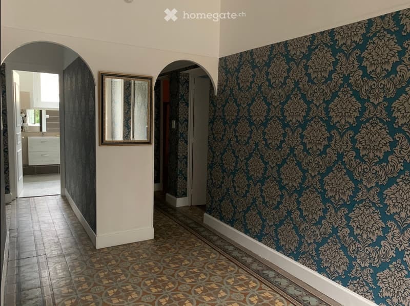 Corridor with patterned wallpaper, decorative mirror, and tiled floor leading to adjacent rooms.