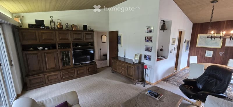 A spacious living room with a beige carpet, wooden cabinetry, a large TV, a sofa, a coffee table, and a dining area with chairs and a table.