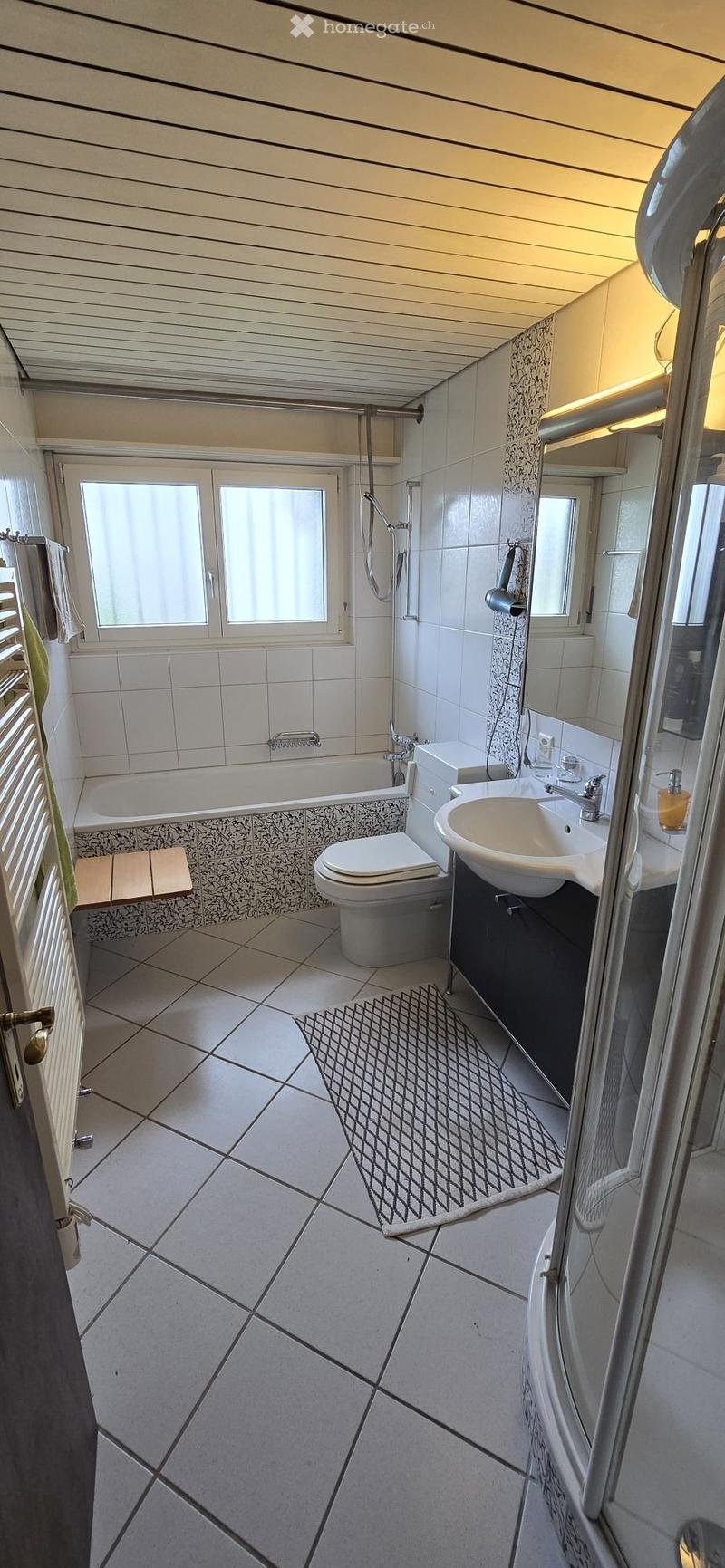 Modern bathroom with white tiles, a bathtub, toilet, sink, mirror, and a shower. Features a patterned rug and a window with blinds.