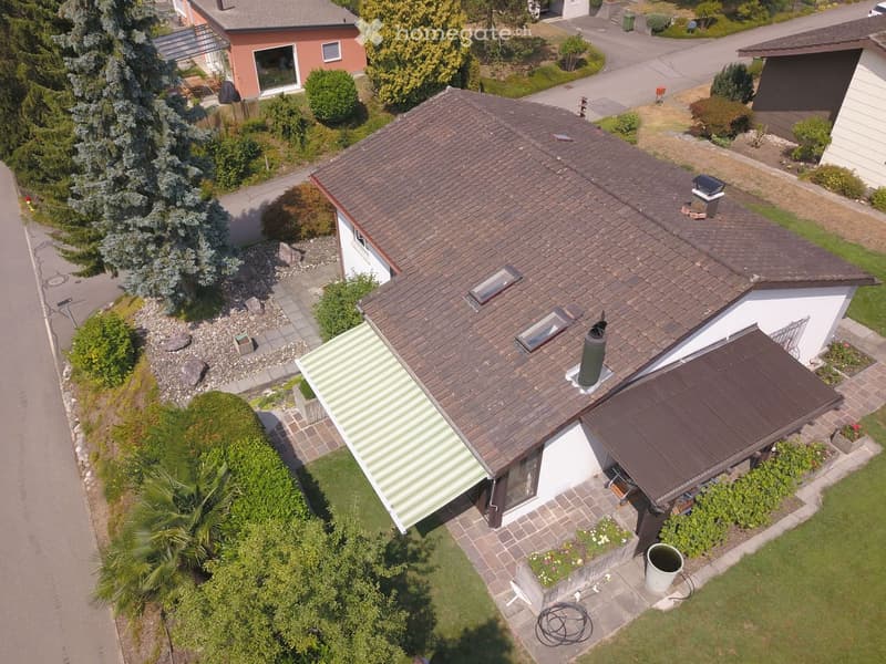Aerial view of a two-story house with a brown tiled roof, white walls, and a well-maintained garden with various plants and trees.