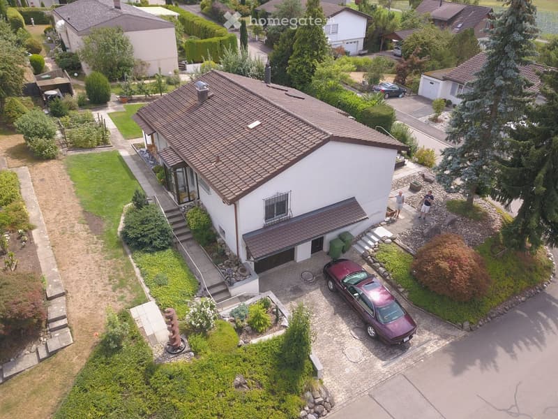 Aerial view of a two-story house with a tiled roof, surrounded by a well-maintained garden and driveway. The house features a garage, a front porch, and is situated in a residential neighborhood with other houses and greenery.