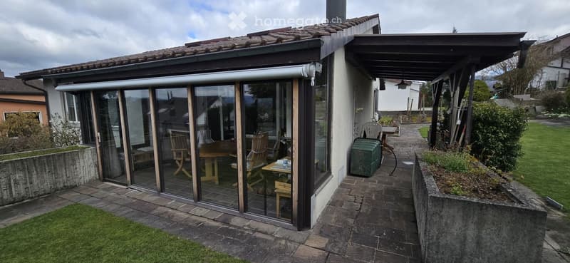 Outdoor patio area with a covered section, glass sliding doors leading to a wooden deck, outdoor furniture including chairs and a table, and a small garden with grass and plants.