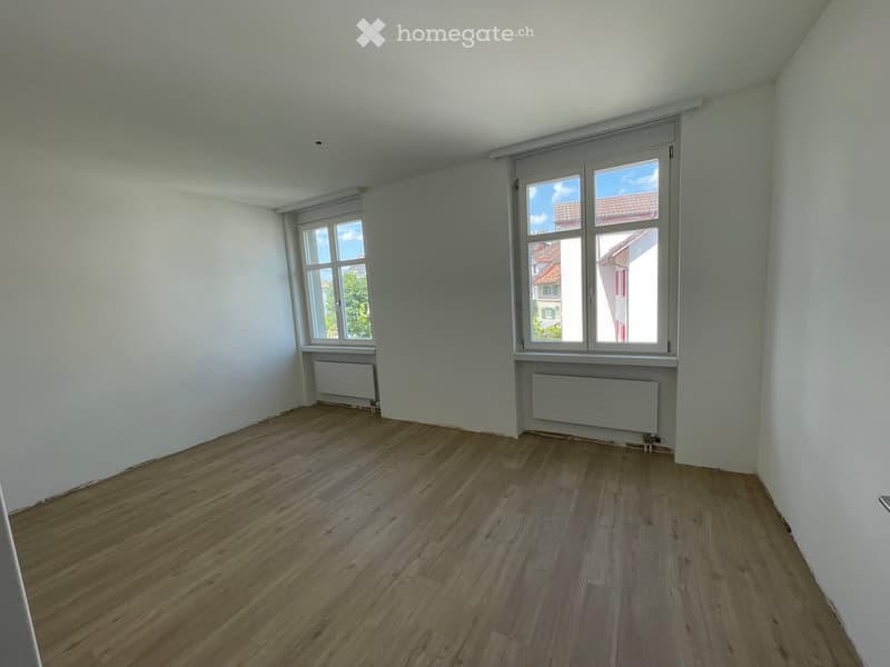 Empty room with wooden flooring, white walls, and two windows offering a view of neighboring buildings.