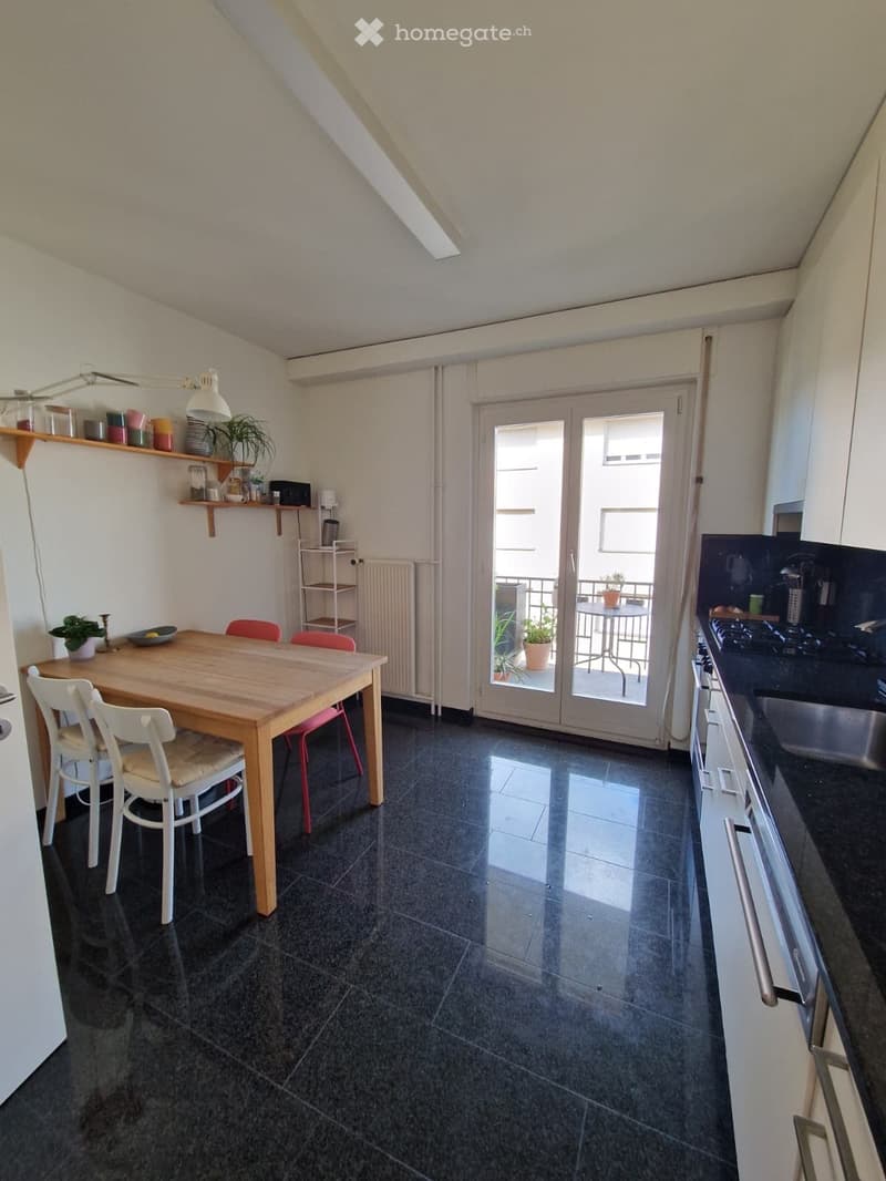Bright, open-plan kitchen-dining area with dark tiled flooring, white walls, and a large sliding glass door leading to a balcony. The space features a wooden dining table with four chairs, a kitchen counter with a sink and faucet, built-in cabinets, a she