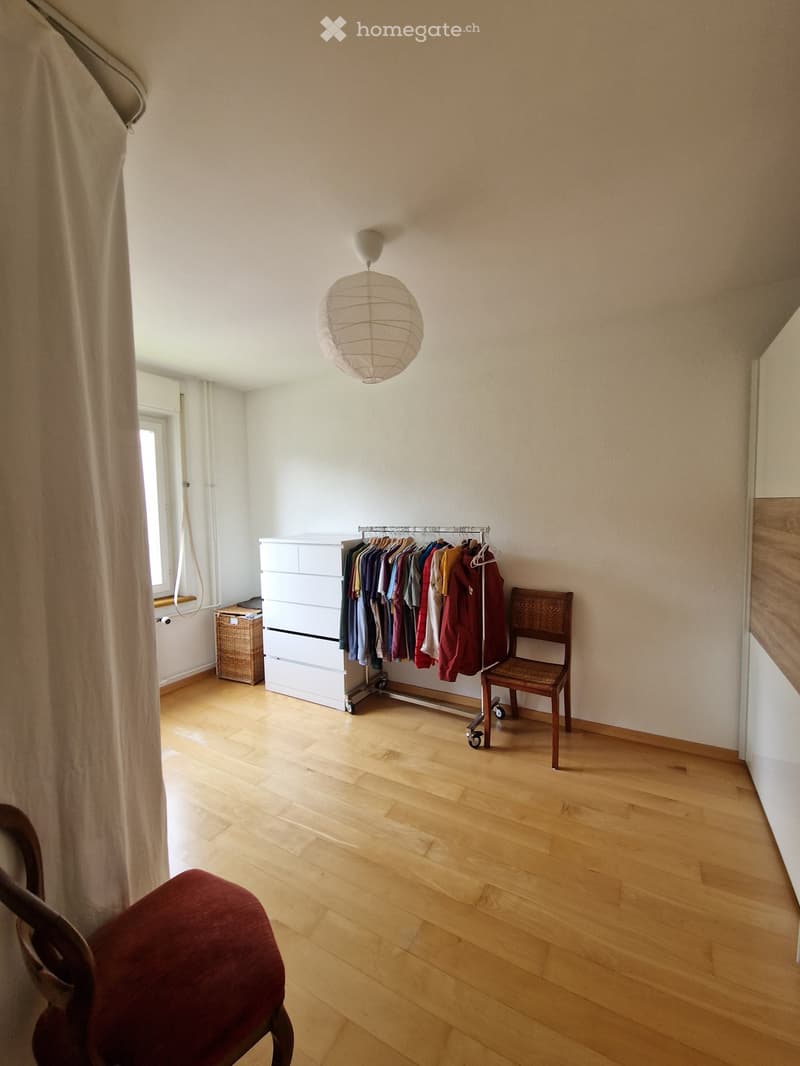A furnished room with wooden flooring, a white wardrobe, a clothing rack with various garments, a wicker basket, and a pendant light fixture.