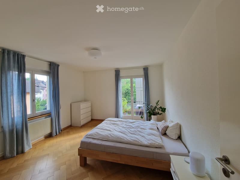 Cozy bedroom with wooden flooring, large windows, a neatly made bed, a nightstand, a drawer, and a potted plant near the window.