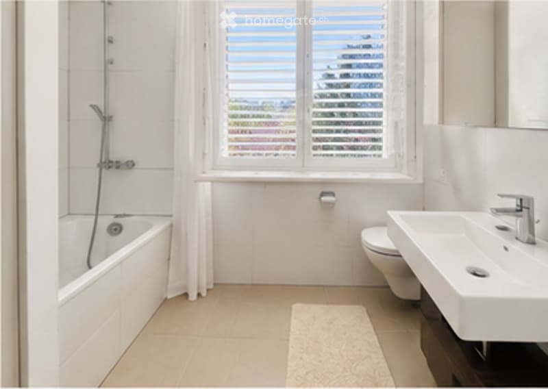 Modern bathroom with a bathtub, toilet, sink, and a window with blinds. The space features white tiles and a light-colored floor.
