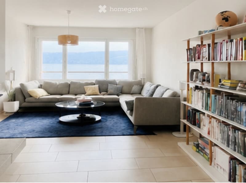 Bright living room with large windows offering a scenic view, featuring a grey sectional sofa, coffee table, bookshelf, and floor lamp.