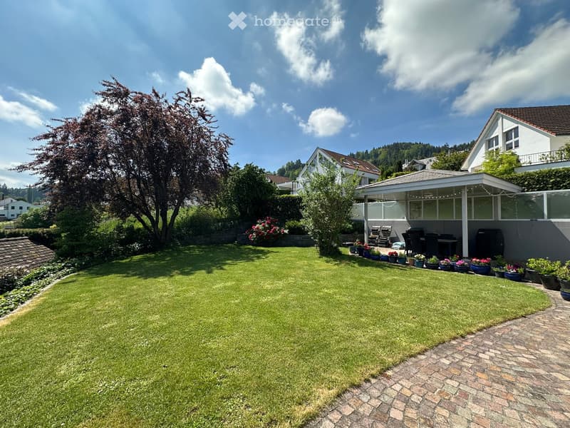 Well-maintained garden with a large lawn, flower pots, patio furniture, and a covered outdoor seating area attached to a house.