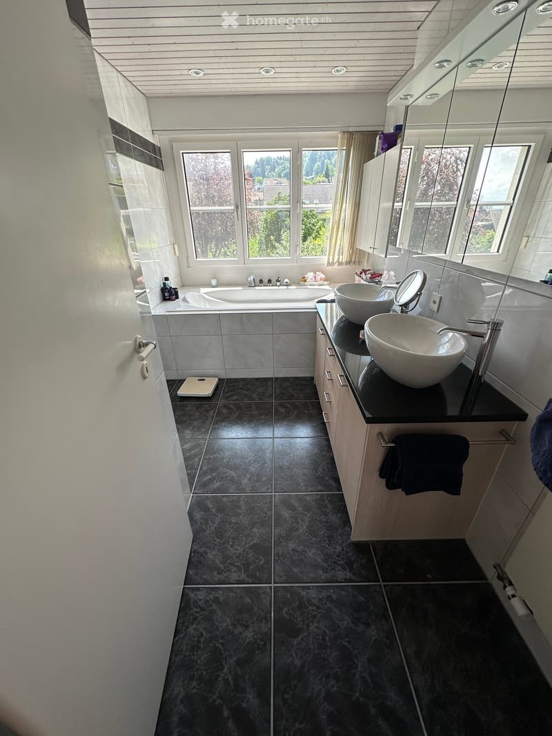 Modern bathroom with a bathtub, two sinks, a large mirror, and a towel hanging on a rack. The room features dark tiled flooring and white walls.