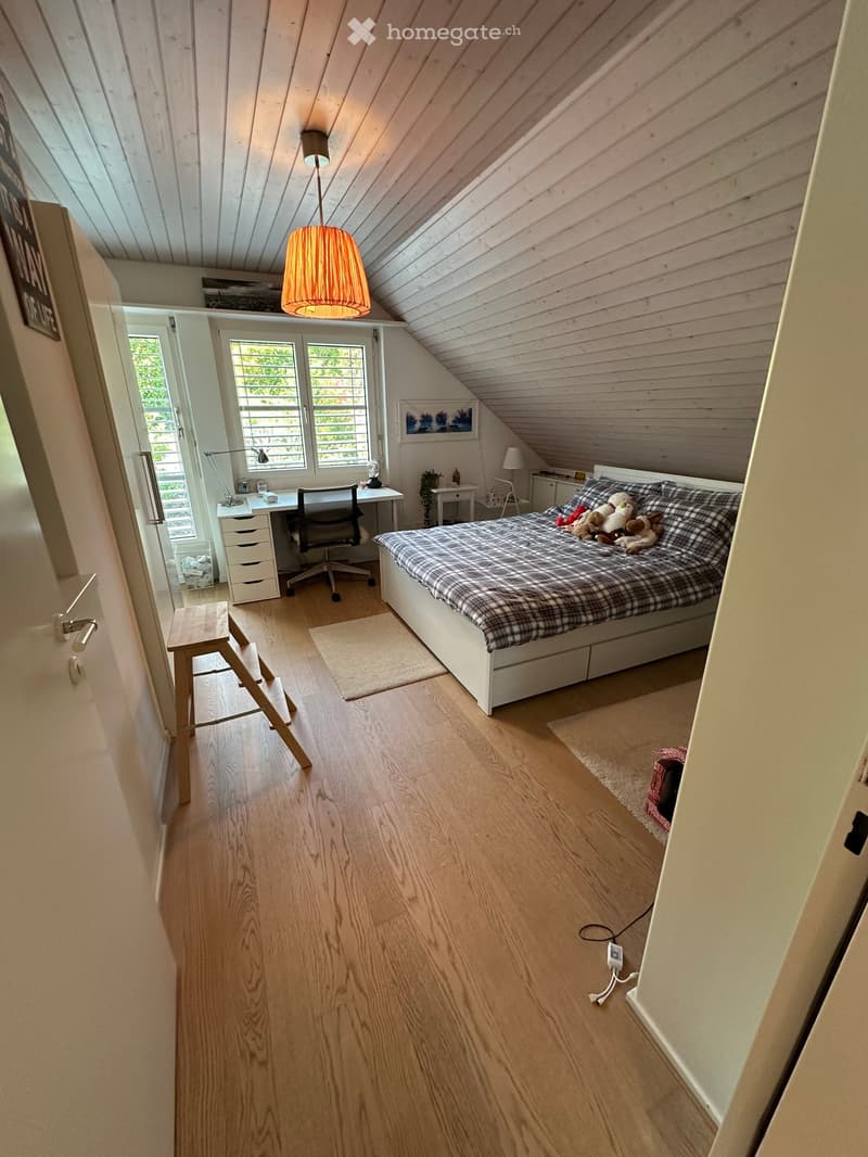 Cozy attic bedroom with wooden floors, slanted wooden ceiling, large windows, bed with plaid blanket, desk with chair, small side table, and pendant lamp.