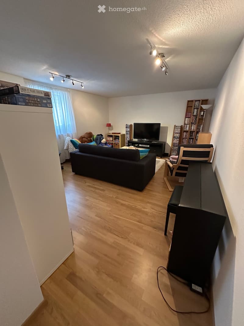Living room with wooden flooring, a black sofa, a piano, a television, a bookshelf, and a small table with a lamp.