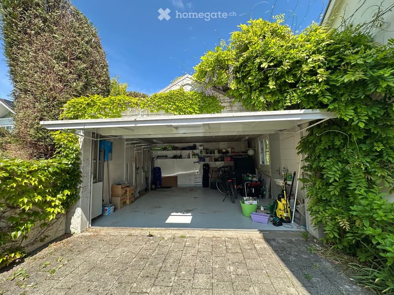 {"type": "Garage", "objects": [], "description": "Open garage with various stored items including bicycles, boxes, and tools, surrounded by lush green foliage and a paved concrete area.", "localized_descriptions": {"EN": "Open garage with various stored i