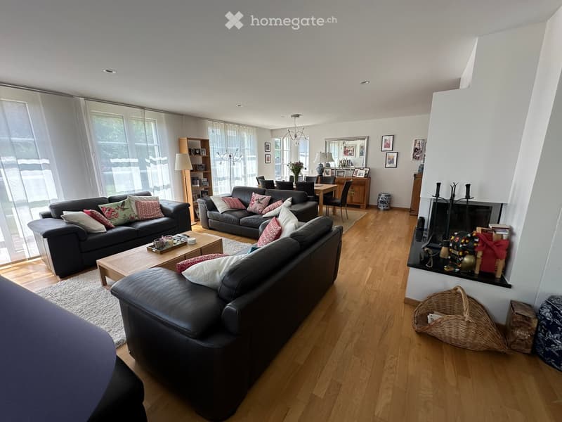 Modern living room with hardwood floors, large windows, three black leather sofas with colorful cushions, coffee table, dining area with chairs and table, fireplace with decorative items, and a wicker basket on the floor.