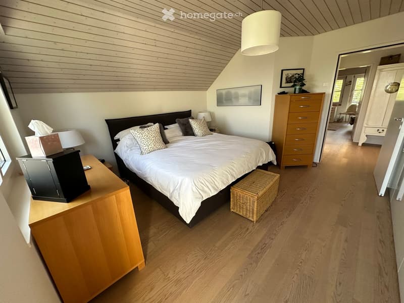 Cozy bedroom with wooden floors, slanted wooden ceiling, large bed with white bedding and patterned pillows, nightstand with lamp, drawer unit, wicker basket, and mirror.