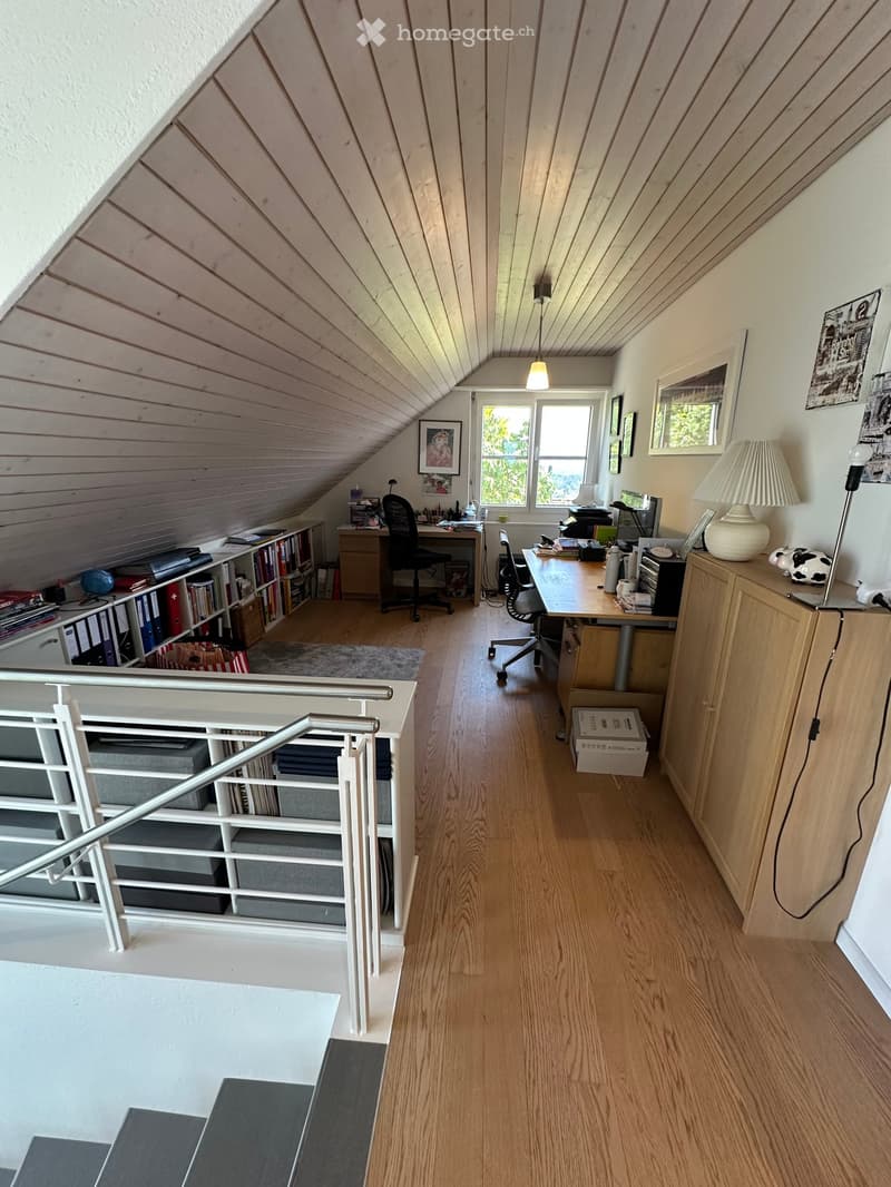 Spacious attic room with wooden walls and slanted ceilings, featuring two desks, chairs, a bookshelf, and various office equipment.