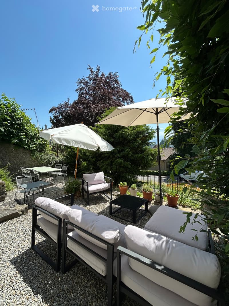 A well-maintained outdoor patio with a sofa, chairs, tables, umbrellas, and potted plants, surrounded by greenery and trees.