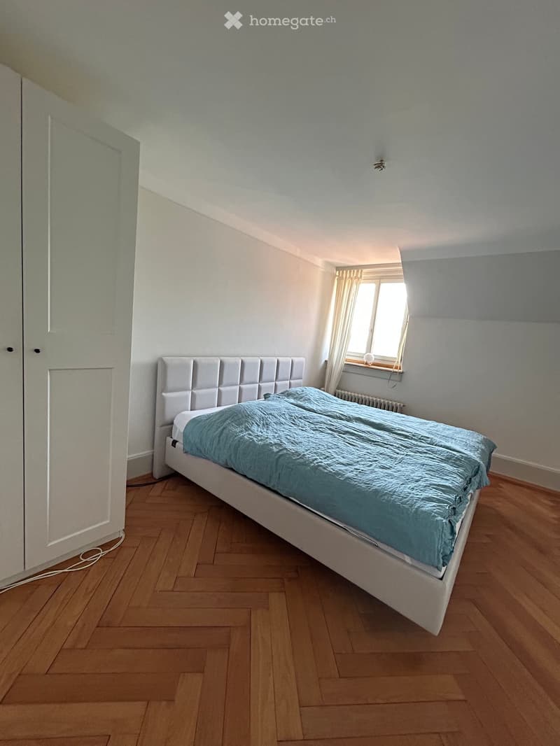 Bedroom with a white bed, headboard, and blue duvet on a wooden floor, with a window and radiator visible.