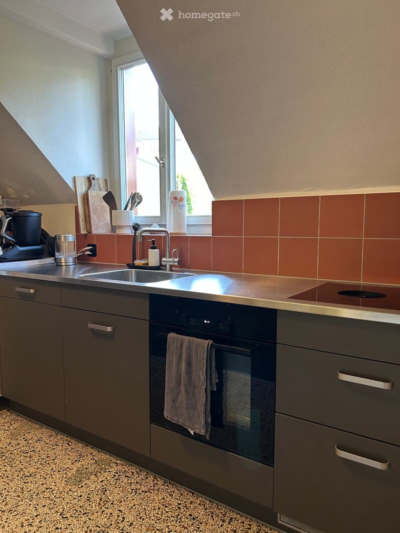 Modern kitchen with stainless steel sink, dark grey cabinets, built-in oven, and a window with a view. Includes a coffee maker, cutting board, and various utensils on the counter.