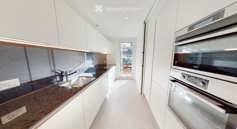 Modern kitchen with white cabinets, dark granite countertops, and stainless steel appliances including a sink and oven. The space is bright with natural light coming through a glass door leading to a balcony.