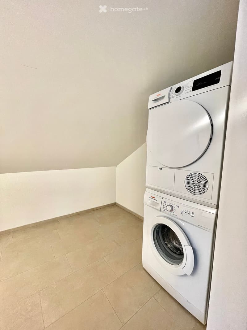 Empty room with beige tiled floor and white walls, featuring a stacked washer and dryer unit in the corner.