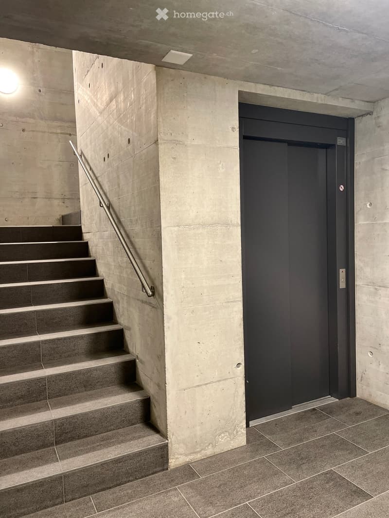 Modern concrete staircase with metal handrail leading to a closed elevator door, featuring tiled flooring and recessed lighting.