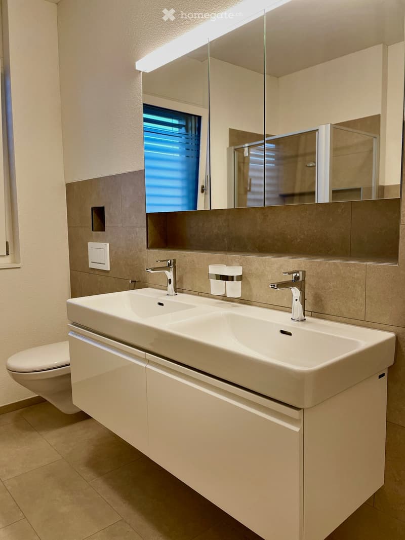 Modern bathroom with a double sink, sleek faucets, a large mirror, and a toilet. The sink has two faucets and two cups placed on it. The walls are tiled in a neutral color scheme, and the floor is tiled as well.