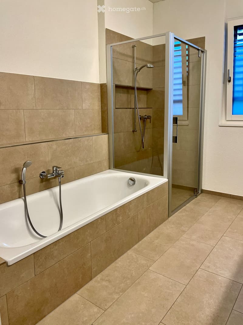 Modern bathroom with a white bathtub, glass-enclosed shower, beige tiles, and a window with blinds.