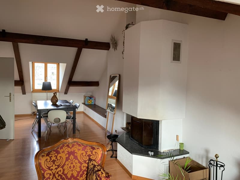 A cozy attic room with wooden beams, a fireplace, a dining table with chairs, a mirror, and a decorative chair near the window.
