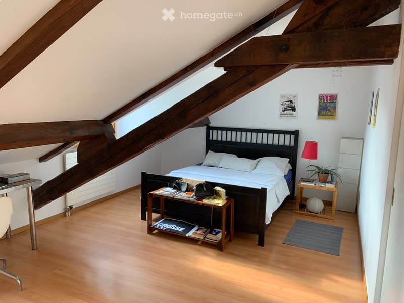 Cozy attic bedroom with wooden beams, slanted ceiling, and large windows. Features a double bed with white linens, bedside tables, a small desk, and a chair.