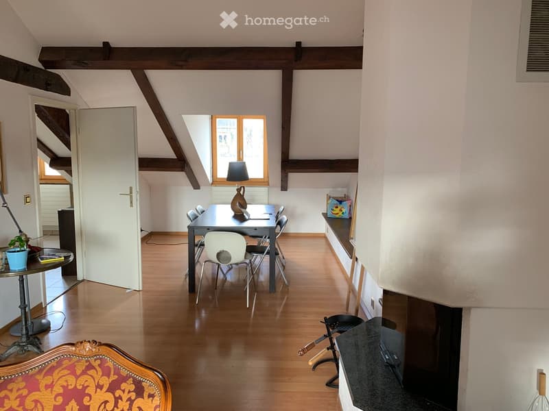 Bright, open room with wooden flooring, exposed wooden beams, and large windows. Features a dining table with chairs, a side table with a lamp and potted plant, and a decorative chair in the foreground.