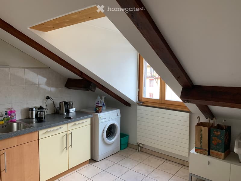 Compact kitchen with wooden cabinets, a sink, a washing machine, and various small appliances on the counter. The room features exposed wooden beams and a skylight window.