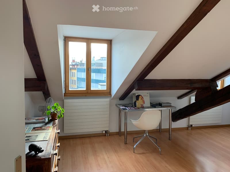 Bright attic room with wooden floors, slanted ceilings, and large windows offering city views. Features a modern desk with a white chair, a small fan, a potted plant, and bookshelves.