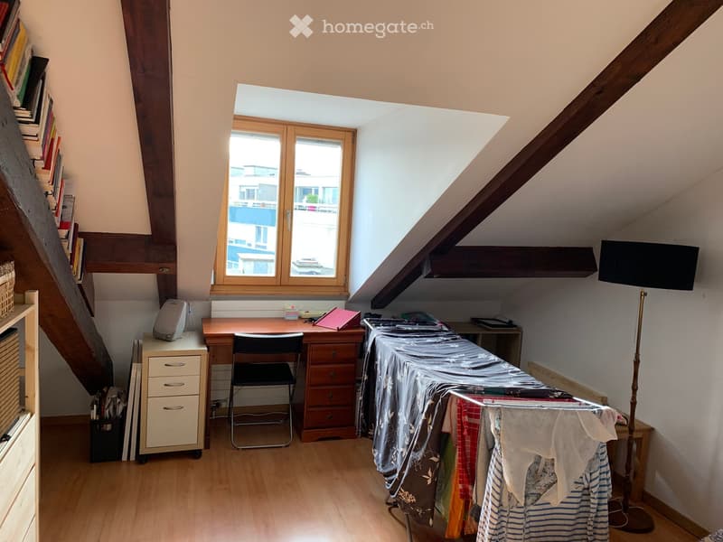 Attic room with wooden floors, slanted ceilings, large windows, desk, chair, cabinet, floor lamp, bookshelf, ironing board, and clothing rack.
