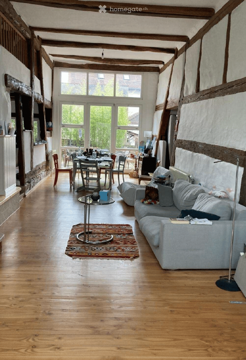Bright, open-plan living and dining area with exposed wooden beams, hardwood flooring, large windows, a grey sofa, coffee table, dining table with chairs, and a bookshelf.