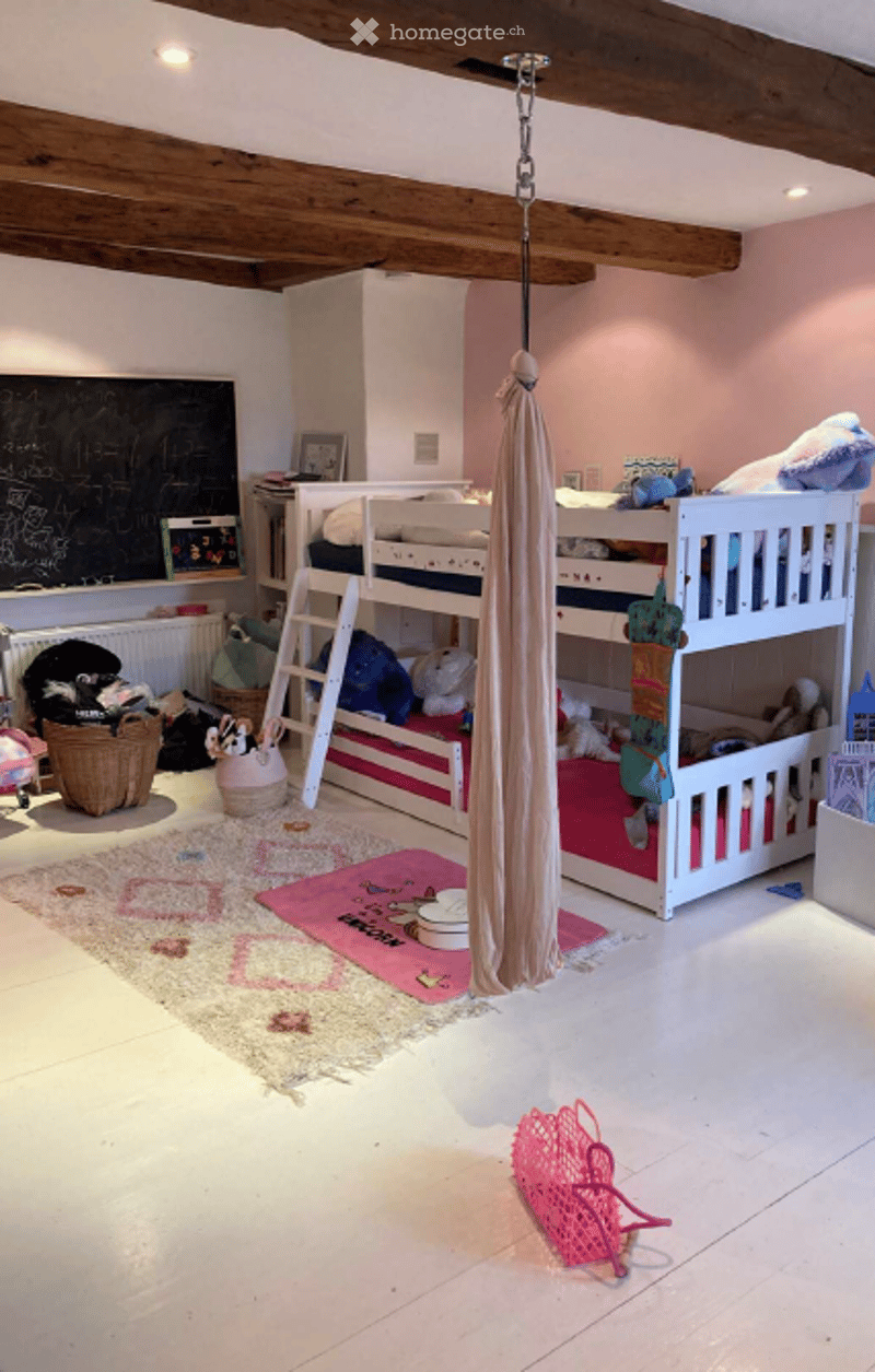 A cozy children's bedroom with a loft bed, colorful toys, a hanging fabric swing, and a chalkboard wall. The room features exposed wooden beams, a white tiled floor, and a soft, pink and cream color scheme.