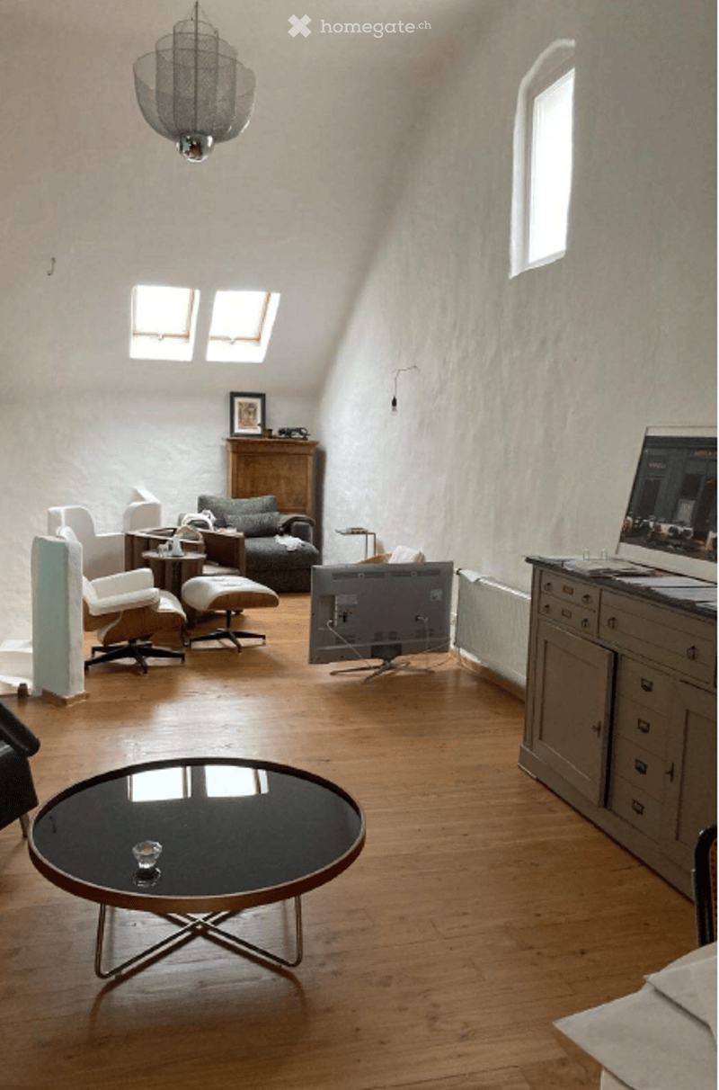 A cozy living room with wooden flooring, a large window, and skylights. Features include a sofa, coffee table, chairs, ottoman, a TV screen, and a cabinet.