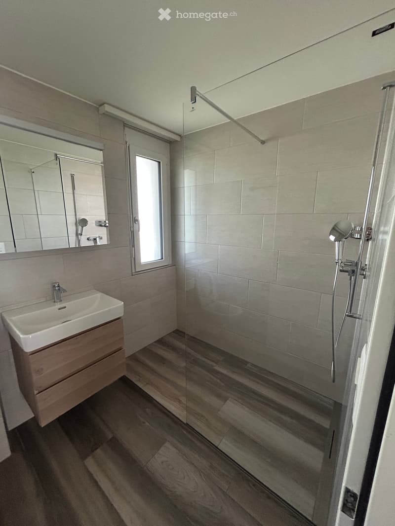 Bathroom with wooden floor, white tiles, glass shower, white sink, wooden cabinet, mirror, and window