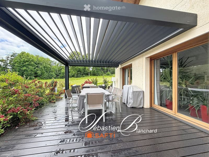 Wooden deck with a covered area, dining table and chairs, grill, and lush greenery in the background.