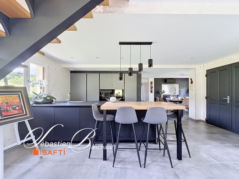 Modern kitchen with a central island, bar stools, pendant lights, and a dining area with a table and chairs. The space features grey cabinets, a grey countertop, and a large window with a view of greenery.