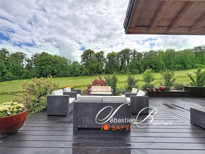 Wooden deck with outdoor furniture including sofas, chairs, and a table, surrounded by greenery and a large open field.