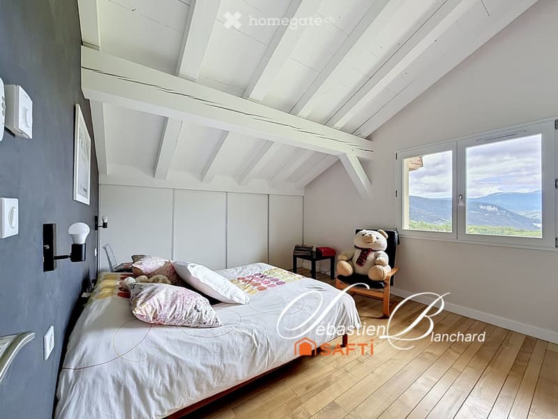 Bright bedroom with wooden flooring, large windows offering a scenic view, a neatly made bed with colorful pillows, a wooden chair with a teddy bear, a small table, and a wall-mounted lamp.