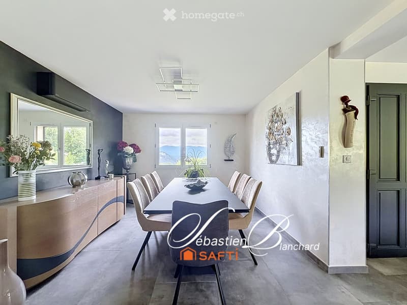 Modern dining room with a long dining table surrounded by chairs, a sleek cabinet, decorative vases, and artwork on the walls. Large windows offer a scenic mountain view.