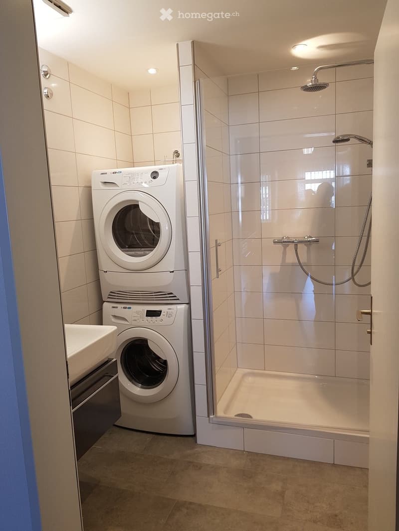 modern bathroom with white tiles, glass shower, white sink, white cabinets, stacked washing machine and dryer