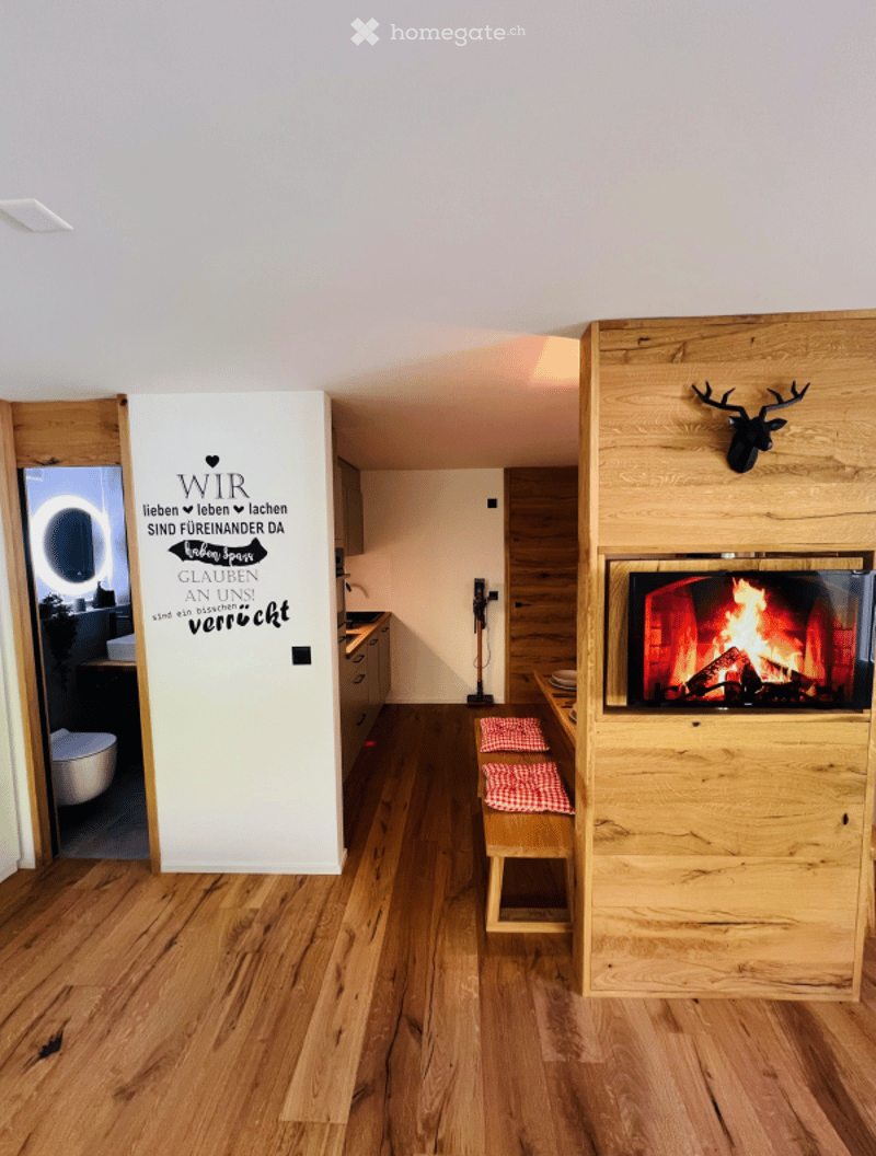 Cozy living space with a modern fireplace, wooden flooring, a bench with checkered cushions, and a decorative mirror. A small toilet is visible in the background.